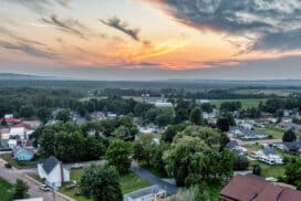 Aerial drone view of a peaceful rural American community village, traditional houses, red rooftops, and straight streets surrounded by lush greenery and open farmland.